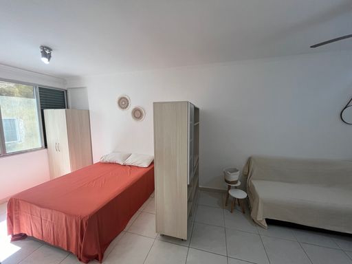 Sala living bem iluminada com uma cama de casal, sofá, mesa e ventilador de teto.