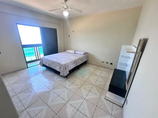 Suíte ampla com muita iluminação natural, ventilador de teto e sacada envidraçada com vista para o mar.