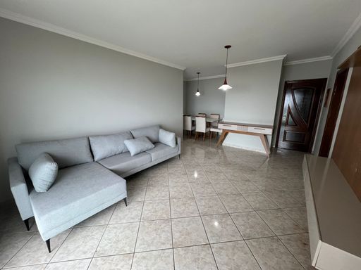 Sala de dois ambientes com painel de TV, sofá cinza com sacada com vista para o mar.