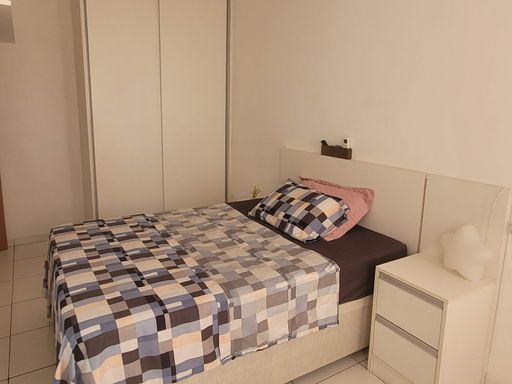 Suíte com guarda-roupa, cama de casal e criado mudo branco.